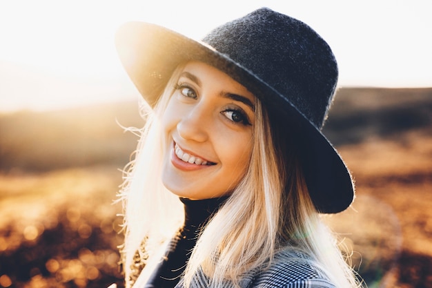 Bardzo młoda kobieta w stylowy kapelusz wesoło uśmiechając się i patrząc na kamery, stojąc na niewyraźne tło jesiennej przyrody w słoneczny dzień. Wesoła kobieta na wsi