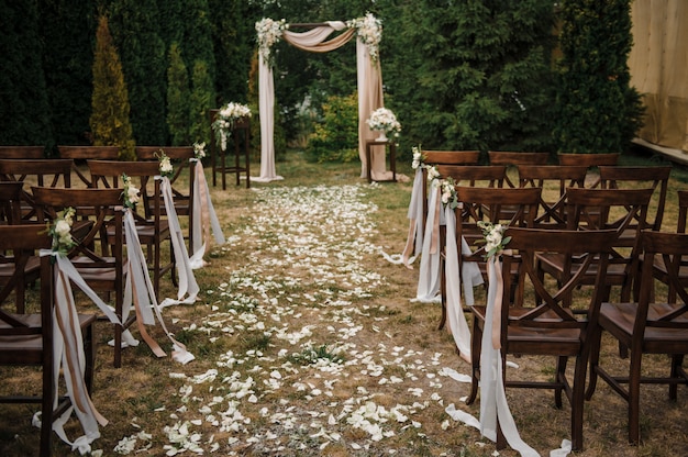 Bardzo piękna ceremonia zewnętrzna. klasyczny ślub w lesie. Łuk jest drewniany. Białe kwiaty. Brązowe krzesła. Obrzęd. Panna młoda i pan młody Dekoracje. Florystyka. Na świeżym powietrzu. Ścieżka z płatków róż.