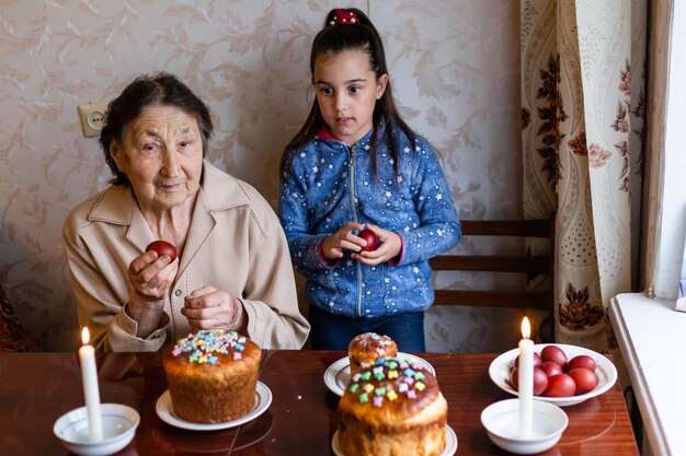 bardzo stara babcia i wnuczka kolorują jajka, przygotowując ciasto wielkanocne na Wielkanoc w domu.