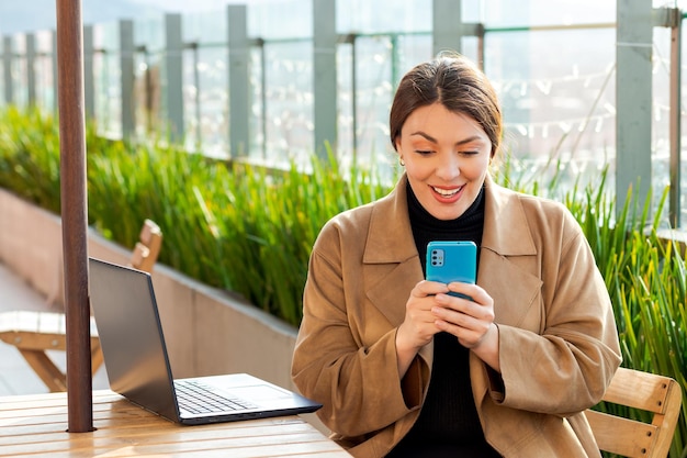 Bardzo szczęśliwa kobieta sprawdza telefon komórkowy z laptopem z dużym uśmiechem na stole obok niej w miejscu pracy na świeżym powietrzu w słoneczne popołudnie