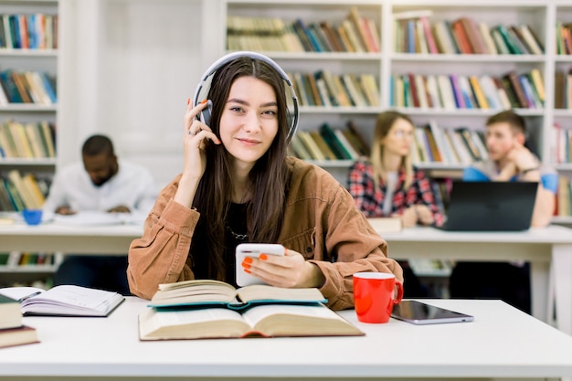 Bardzo Szczęśliwa Uśmiechnięta Młoda Studentka W Modnych Ubraniach, Siedząca W Bibliotece I Słuchająca Dobrej Muzyki W Słuchawkach,