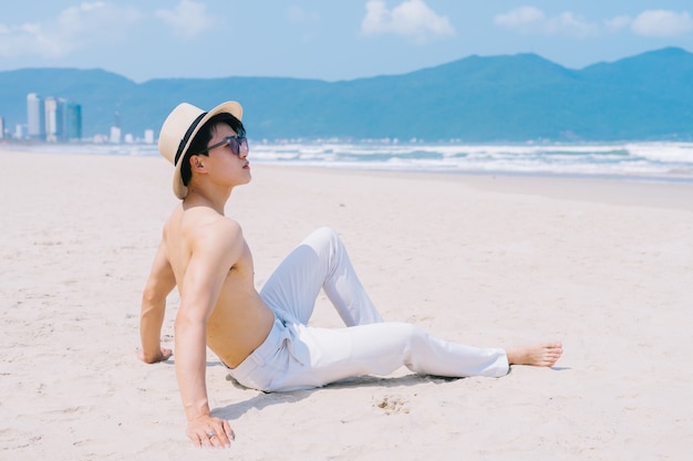 Barebacked młody człowiek Azji siedzi na piasku i patrząc na morze