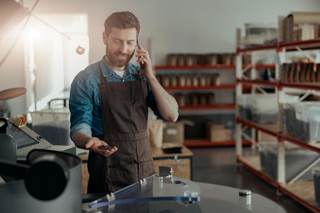 Barista rozmawia przez telefon i patrzy na pieczone ziarna kawy na tle maszyny do pieczenia