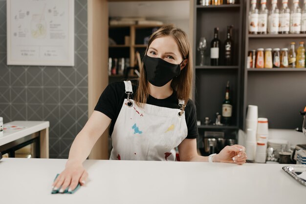 Barista w czarnej masce medycznej uśmiecha się i czyści blat barowy w kawiarni. Ładna kelnerka dezynfekująca kawiarnię.