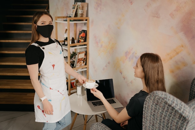 Barista w czarnej masce na twarzy serwujący dziewczynie latte w kawiarni