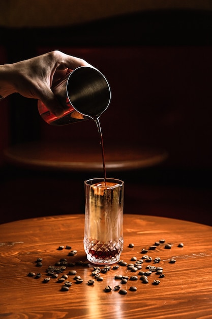 Barman nalewający koktajl z shakera do szklanki typu highball collins z włócznią lodową. Ziarna kawy na drewnianym stole.