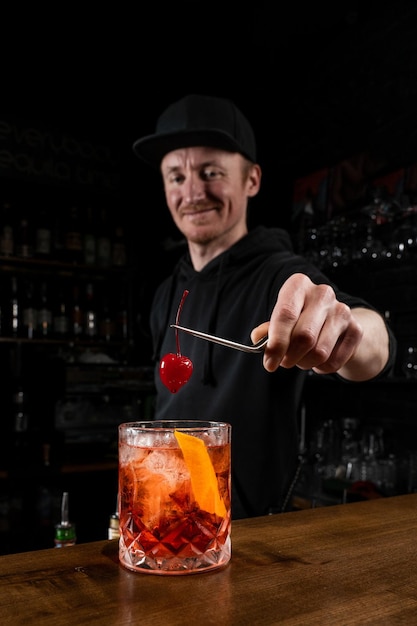 Barman trzyma wiśnię nad koktajlem Negroni w szkle Barman przygotowuje klasyczny koktajl Negroni w barze