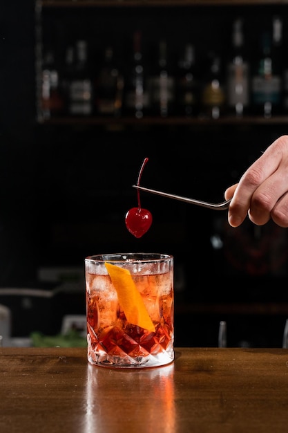 Barman trzyma wiśnię nad koktajlem Negroni w szkle Barman przygotowuje klasyczny koktajl Negroni w barze