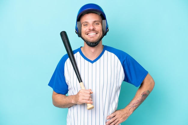 Baseballista z kaskiem i kijem odizolowywającym na błękitnym tle pozuje z rękami przy biodrem i ono uśmiecha się