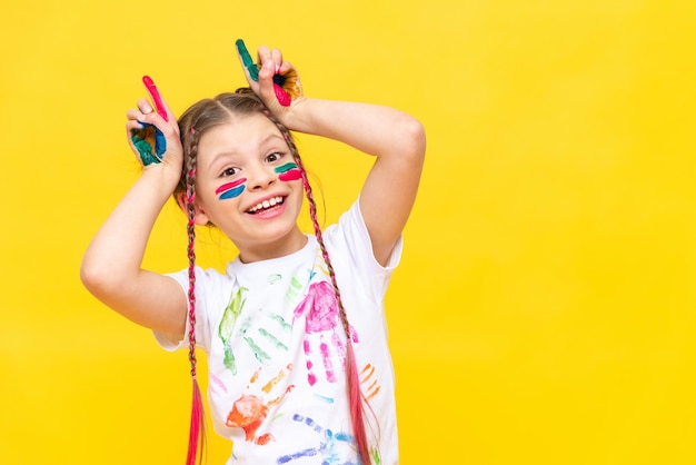 Bawiąca się dziewczyna poplamiona farbami pokazuje rogi na żółtym, odizolowanym tle Dziecko bawi się kolorowymi farbami Kursy rozwoju kreatywności dzieci Wesołe gry z dzieciństwa