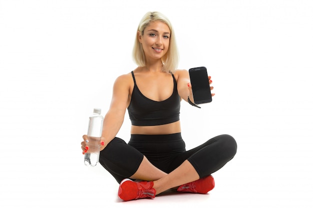 Beauiful sportowa kaukaska dziewczyna siedzi na podłodze w res tenisówkach, krzyżuje nogi, pokazuje telefon komórkowy i trzyma butelkę wody w izolacji