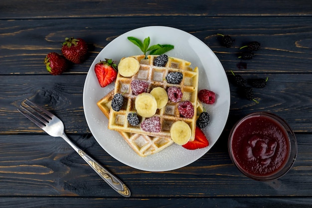 Belgijskie gofry z letnimi jagodami i cukrem pudrem w białym talerzu na ciemnym drewnianym tle Słodkie belgijskie gofry na śniadanie lub lunch