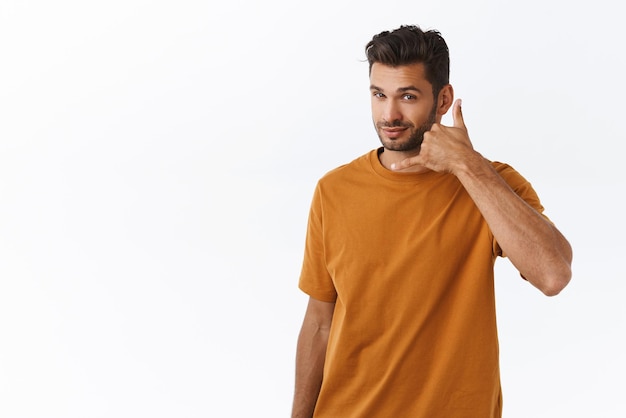 Bezczelny romantyczny seksowny brodaty mężczyzna w brązowej koszulce proszący o telefon uśmiechnięty chytry i bezczelny wykonujący gest telefoniczny ręką w pobliżu ucha, zachęcający do pójścia na randkę z nim stojąc na białym tle