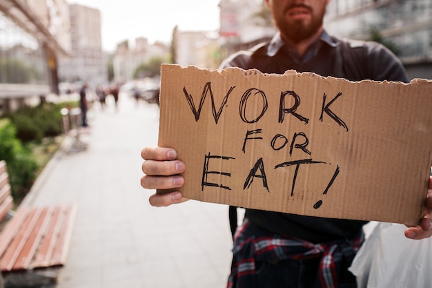 Bezdomny stojący na zewnątrz i trzymający karton z napisem praca do jedzenia