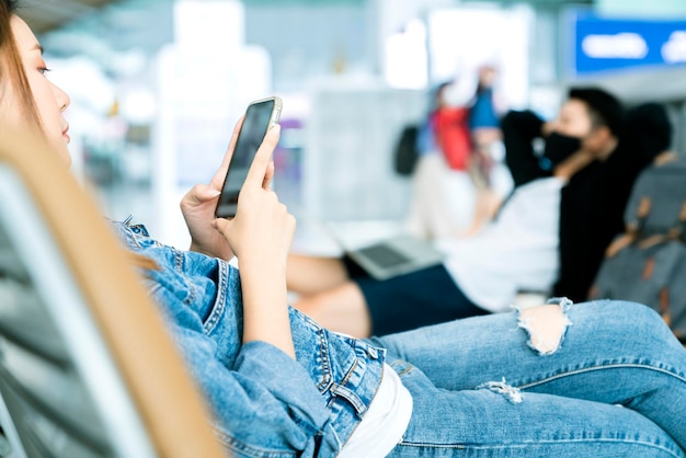 Bezpieczna podróż azjatycka para podróżnik nosi maskę ochronną na twarz czekaj zrelaksuj się z dystansem społecznym nowy styl życia na lotnisku terminal odlotów używaj smartfona i laptopa do harmonogramu
