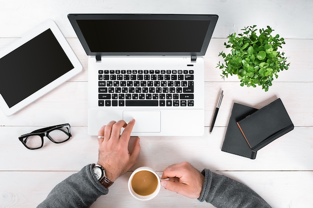 Bezpośrednio nad widokiem ludzkich rąk piszących na laptopie. Laptop, cyfrowy tablet, pamiętnik, filiżanka kawy i roślina doniczkowa na biurku. Człowiek pracujący w domu.