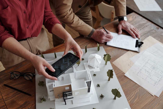 Zdjęcie bezpośrednio nad widokiem nierozpoznawalnego asystenta fotografującego model 3d domu, podczas gdy architekt robi notatki o przestrzeni pokoju