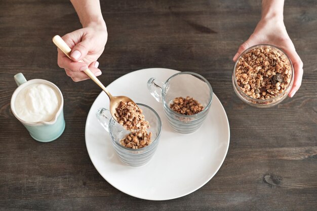 Bezpośrednio powyżej widok nierozpoznawalnej kobiety wkładającej musli do kubka mieszając je z jogurtem na muesli