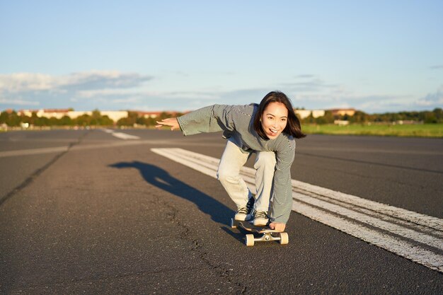 Zdjęcie beztroska łyżwiarka na deskorolce jeżdżąca na longboardzie na pustej drodze trzymająca się za ręce bokiem i