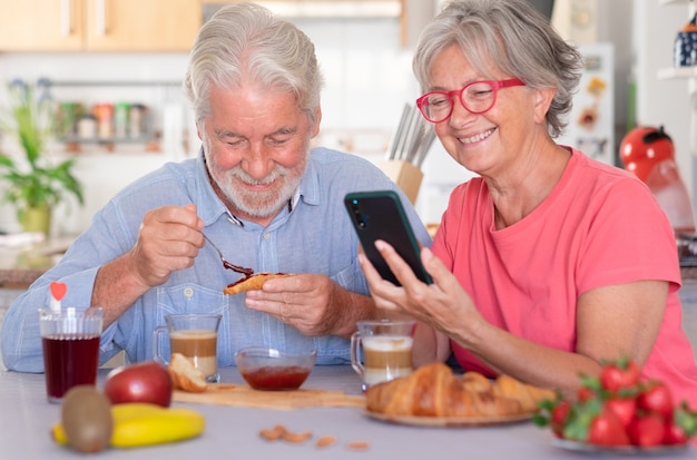 Beztroska para kaukaski jedząca śniadanie w domu. Żona za pomocą telefonu komórkowego. Seniorzy zrelaksowani i szczęśliwi, cieszący się jedzeniem i piciem