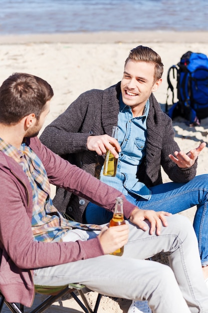 Beztroska rozmowa. Widok z góry dwóch przystojnych młodych mężczyzn pijących piwo i rozmawiających ze sobą, siedząc razem na plaży