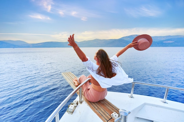 Beztroska, zadowolona, szczęśliwa, inspirowana podróżniczka z otwartymi ramionami cieszy się relaksującymi wakacjami na białej prywatnej łodzi na turkusowym morzu
