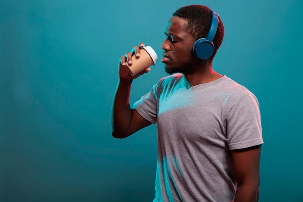 Beztroski facet słuchający muzyki na zestawie słuchawkowym i pijący kawę z kubka na relaks w studio. Pewna dorosła osoba z bezprzewodowymi słuchawkami, ciesząca się napojem i dźwiękiem mp3 na gadżecie audio.