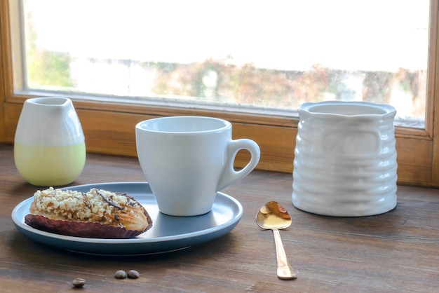 Biała filiżanka kawy espresso, świeży upieczony deser słodki ekler na płycie kuchennego stołu przed oknem, naczynia naczynia, dzbanek do mleka, dom zielonych roślin. poranne francuskie śniadanie domowe, miejsce na kopię