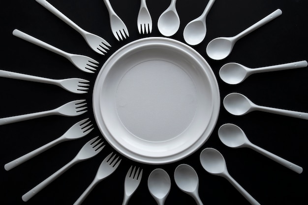 Zdjęcie biała jednorazowa zastawa stołowa, talerz i wokół leżą łyżki i widelce na czarnym tle