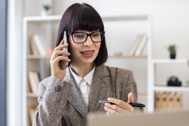 Biała kobieta rozmawiająca przez telefon komórkowy podczas oglądania ekranu laptopa w biurze