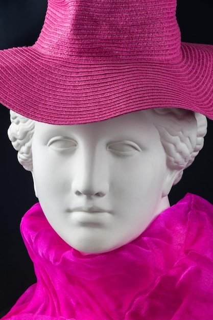 Biała tynkowana kopia antycznego posągu głowy w różowym kapeluszu i szaliku na czarnym tle. Gipsowa rzeźba kobiecej twarzy.