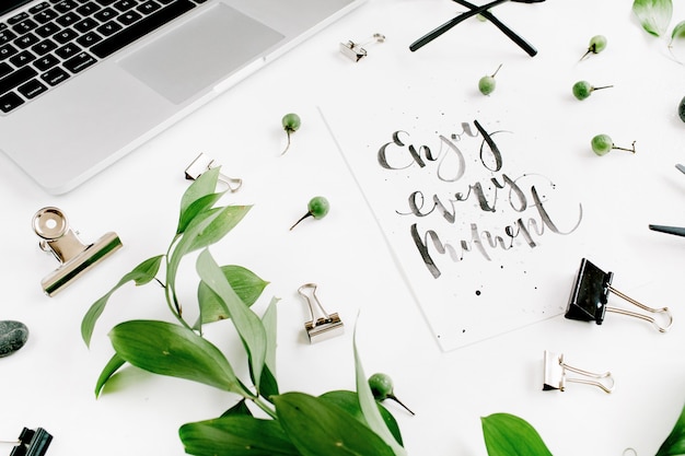 Zdjęcie białe biurko z cytatem enjoy every moment, zielonymi liśćmi i materiałami biurowymi