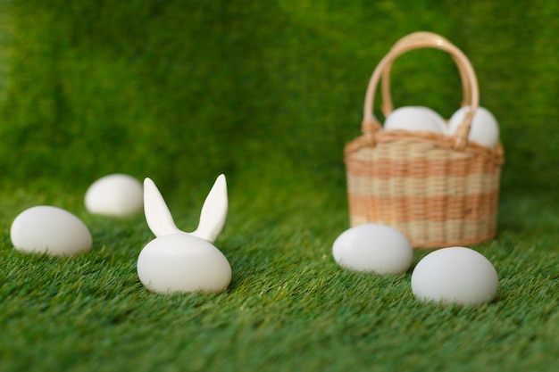 Białe kurze jajka na zielonej trawie za którymi ukryte są uszy wielkanocnego królika Wiklinowy kosz Symbole obchodów świąt religijnych wśród chrześcijan i katolików