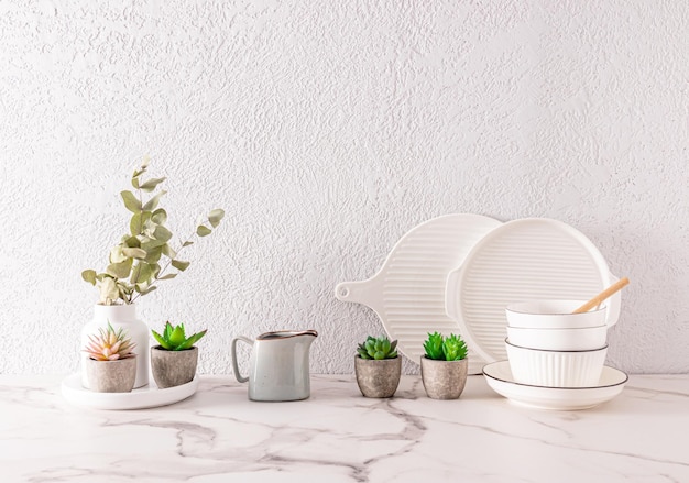 Białe naczynia ceramiczne i kwiaty wewnętrzne w ceramicznych mini doniczkach na blacie kuchennym wykonanym z białej marmurowej szarej ściany cementowej w przestrzeni do kopiowania