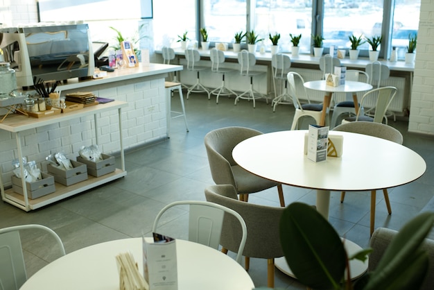 Białe okrągłe stoły otoczone wygodnymi fotelami i krzesłami wzdłuż okna w przytulnej kawiarni