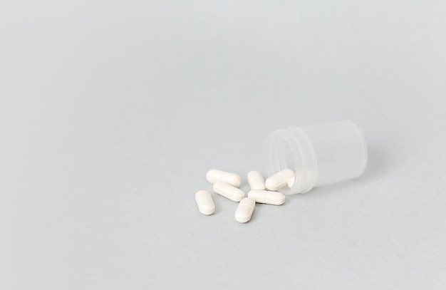 białe tabletki wylewają się z plastikowego pojemnika na szarym tle
