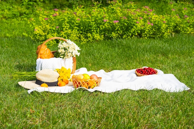 Biały koc piknikowy na świeżej trawie z koszykiem owoców kwiatów słomkowy kapelusz