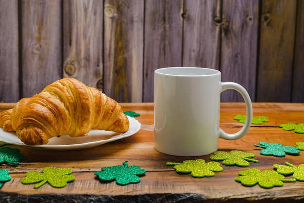 Biały kubek i rogaliki na drewnianym stole z błyszczącymi koniczynami St Patricks Day makieta