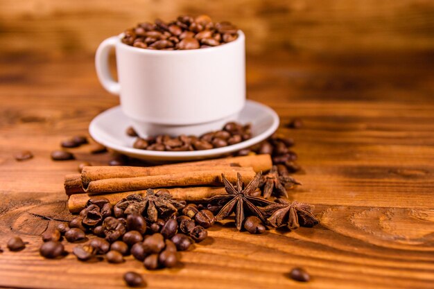 Biały kubek wypełniony ziarnami kawy, anyżem i cynamonem na rustykalnym drewnianym stole