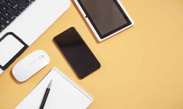 Biały laptop, smartfon, notatnik, długopis, tablet i mysz na żółtym tle.