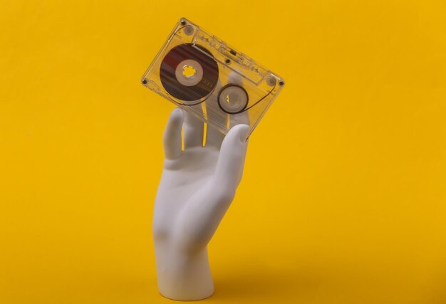 Biały manekin ręki trzymającej retro kaseta audio na żółtym tle.