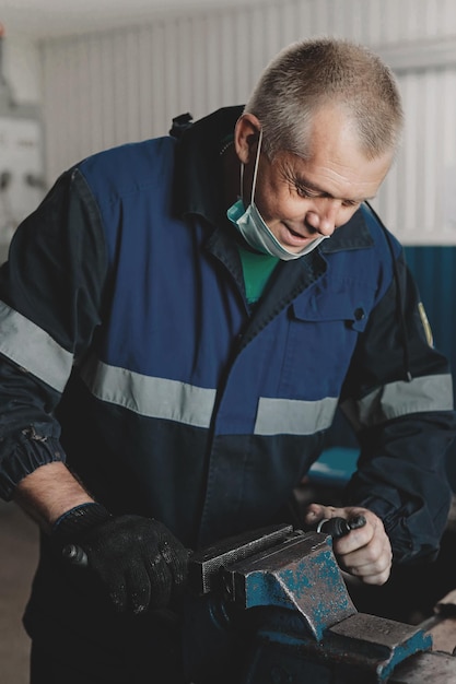 Biały mężczyzna w kombinezonie pracuje w pokoju za stołem warsztatowym Mechanik lub ślusarz zaciska metalową część w imadle