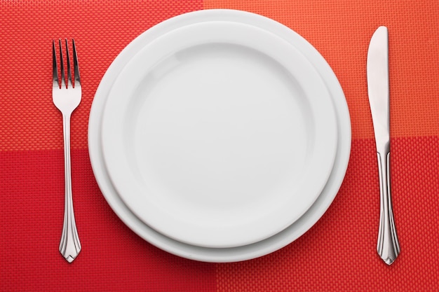 Biały pusty talerz z widelcem i nożem na czerwonym obrusie