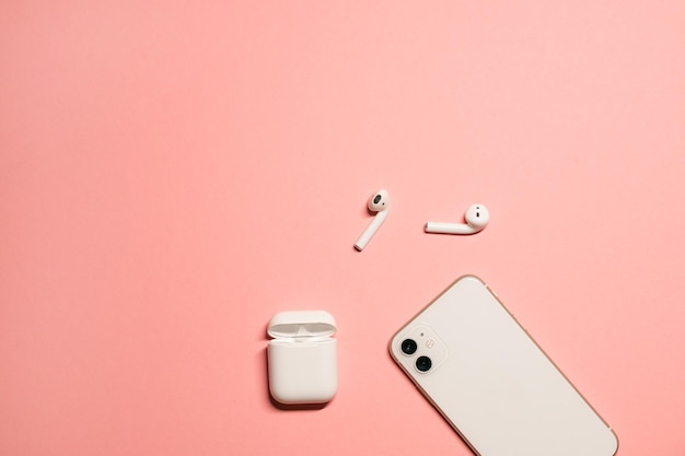 biały smartfon i nowoczesne bezprzewodowe słuchawki z etui ładującym na pastelowym różowym tle