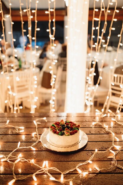 Biały tort weselny na stole ozdobionym świecącymi girlandami