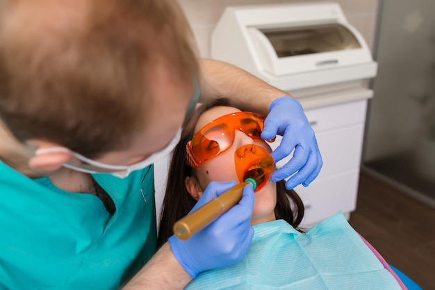 Zdjęcie biedna dziewczyna w śliniaku dla pacjenta i okularach ochronnych w gabinecie stomatologicznym dentysta w niebieskich lateksowych rękawiczkach usuwa ząb za pomocą zwijacza policzkowego i kleszczyków zbliżenie poziome zdjęcie