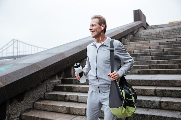 Biegacz w szarym stroju sportowym stojący na schodach z butelką wody i torbą