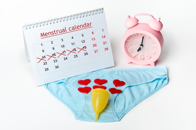 Zdjęcie bielizna damska kubek menstruacyjny z czerwonymi serduszkami budzik i kalendarz menstruacyjny z przekreślonymi datami na białym tle problem zdrowia kobiet kalendarz menstruacyjny dla kobiet