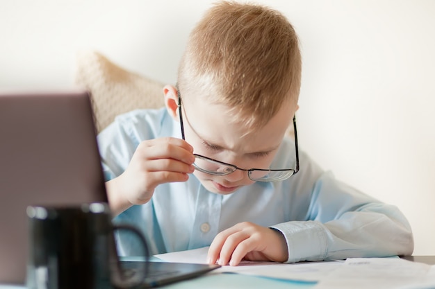 Biurko Workspace z laptopem i filiżanką kawy. Mały śliczny biznesowy chłopiec obsiadanie przy stołem i patrzeć na ważnych dokumentach. Dziecko uczy się online.