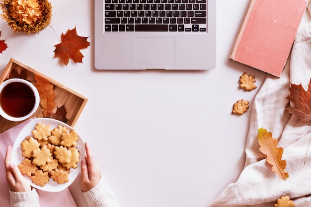 Biurko z laptopem, filiżanką kawy, ciastkami, książką i jesiennymi liśćmi. Widok z góry